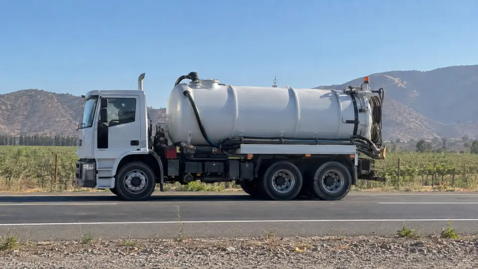 Camión de succión blanco en una carretera rural con viñedos y cerros secos de fondo