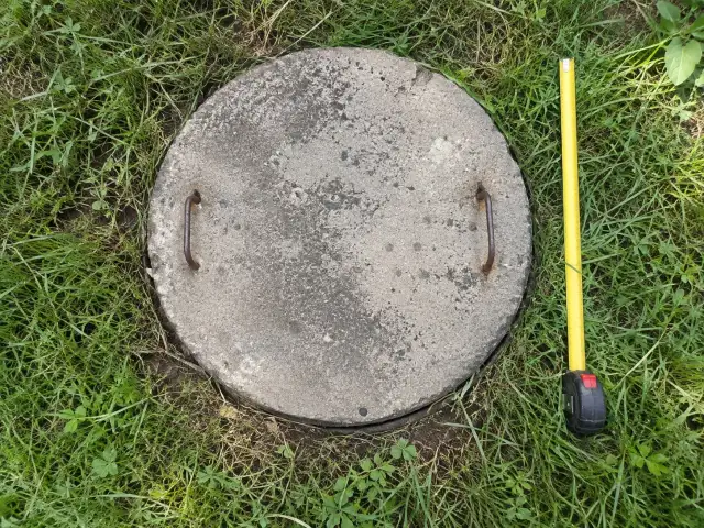 Tapa circular de hormigón de una fosa séptica en el pasto, junto a una huincha de medir